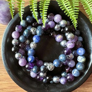 May include: A collection of purple, blue, and silver beaded bracelets with silver accents. The bracelets are arranged in a bowl.