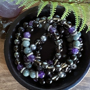 May include: A collection of beaded bracelets in a black bowl. The bracelets feature a mix of black, gray, and purple beads. Some beads are faceted, and others are smooth. The bowl sits on a wooden surface, with a purple crystal and greenery in the background.