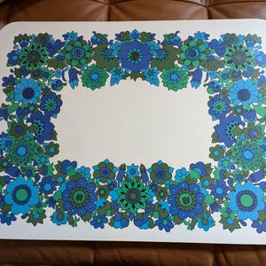 May include: White placemat with a blue and green floral border. The flowers are stylised and have a retro look.