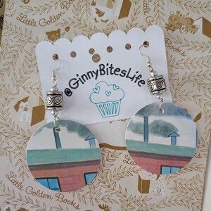 May include: A pair of dangle earrings featuring circular paper cutouts with a printed illustration of a house roofline. Each earring is suspended from a silver-toned metal hook and accented with a decorative, textured silver-toned metal barrel bead.