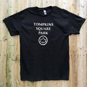 May include: Black t-shirt with white text that reads "Tompkins Square Park" and a white maple leaf logo.