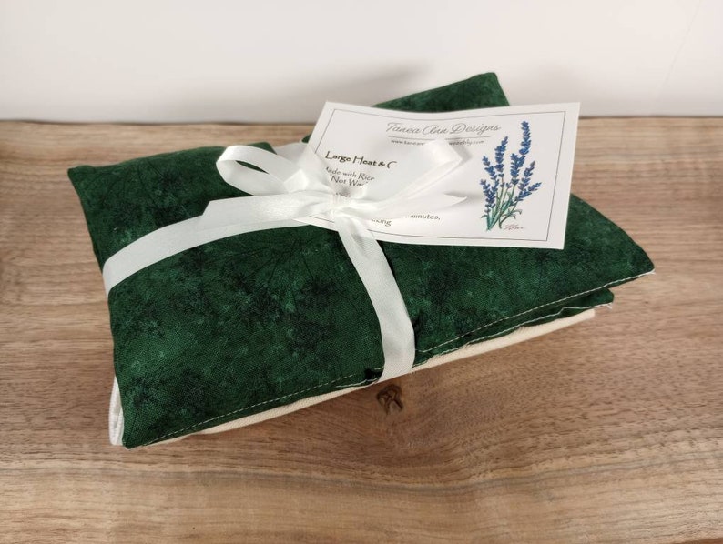 K&ouml;nnte beinhalten: Ein gr&uuml;nes Stoff-W&auml;rmep&auml;ckchen, gebunden mit einem wei&szlig;en Band und einem Etikett mit der Aufschrift "Large Heat & C Made with Flour Rice Wheat" und "Toesera Clean Designs" mit einer Illustration einer Lavendelbl&uuml;te.