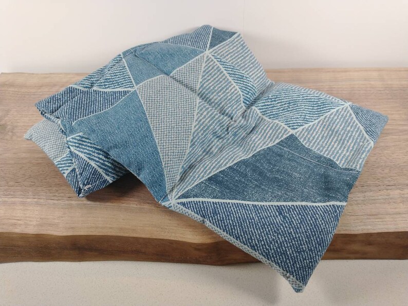 K&ouml;nnte beinhalten: Ein blaues und wei&szlig;es Heizkissen aus Stoff mit geometrischem Muster. Das Kissen ist in der Mitte gefaltet und liegt auf einer Holzoberfl&auml;che.