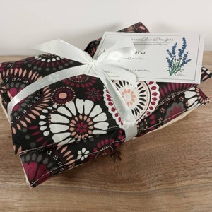 K&ouml;nnte beinhalten: Zwei Lavendel-W&auml;rmepads, die mit einem wei&szlig;en Band umwickelt sind. Die W&auml;rmepads bestehen aus schwarzem Stoff mit einem rosa-wei&szlig;en Blumenmuster. Das Etikett auf dem oberen W&auml;rmepad lautet "Heat Pad" und "Lavender".