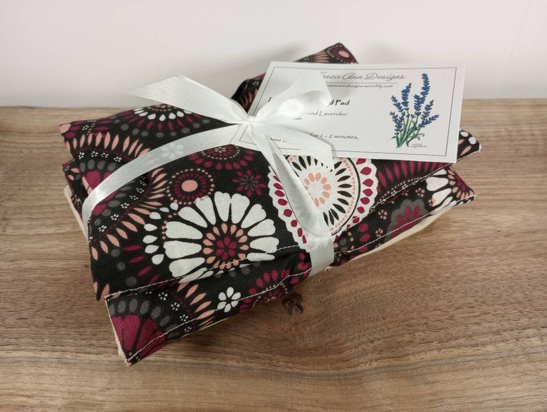 K&ouml;nnte beinhalten: Zwei Lavendel-W&auml;rmepads, die mit einem wei&szlig;en Band umwickelt sind. Die W&auml;rmepads bestehen aus schwarzem Stoff mit einem rosa-wei&szlig;en Blumenmuster. Das Etikett auf dem oberen W&auml;rmepad lautet "Heat Pad" und "Lavender".