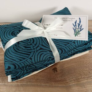 K&ouml;nnte beinhalten: Ein Set aus drei tealblauen und schwarzen W&auml;rmekissen mit Muster, die mit einem wei&szlig;en Band zusammengebunden sind. Die W&auml;rmekissen sind in ein wei&szlig;es Tuch gewickelt und haben einen Rei&szlig;verschluss. Eine kleine Karte mit dem Text "The Clean Designs" ist am Band befestigt.