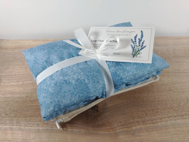 K&ouml;nnte beinhalten: Ein blauer und wei&szlig;er W&auml;rmekissen mit Blumenmuster, das mit einem wei&szlig;en Band umwickelt ist. Das Kissen ist mit "Large Heat Cold Pack Lavender and Rice" beschriftet.