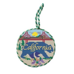 May include: A round, cross-stitched ornament featuring a blue bear silhouette with the word "California" in gold script. The design includes a red bridge, a yellow sun, and green trees, with a green and white twisted cord for hanging.