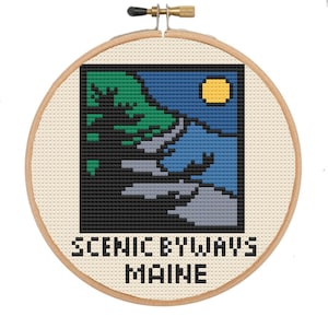 Peut inclure: Un cercle de broderie rond en bois avec un motif de point de croix représentant une scène de montagne avec un soleil jaune. Le texte "Scenic Byways Maine" est situé sous le motif.