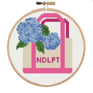 May include: Cross-stitch embroidery in a wooden hoop. The design features a pink tote bag with the letters "NDLPT" and a bouquet of blue hydrangeas with green leaves. The background is white.