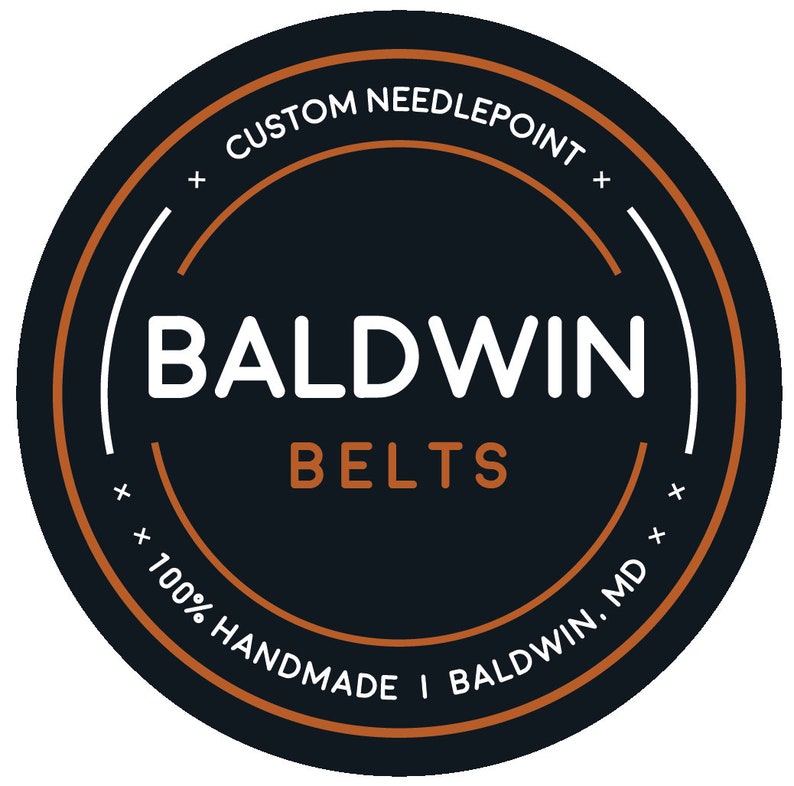 May include: A black and white logo with the text "CUSTOM NEEDLEPOINT" in a circle around the text "BALDWIN BELTS" with the text "100% HANDMADE I BALDWIN. MD" in a circle around the bottom.