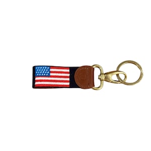 May include: A navy blue key chain with a small American flag woven into the strap. The key chain has a brown leather loop and a gold metal clasp.