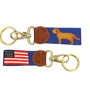 May include: Two keychains with blue and white striped fabric and leather accents. One keychain features a golden retriever dog and the other features an American flag.