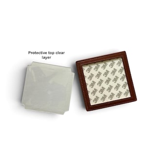 May include: A square object with a brown frame and a white adhesive backing featuring a repeating 3M logo pattern, alongside a separate light gray square piece with a protective top clear layer and text indicating the protective top clear layer.