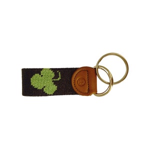 May include: A keychain featuring a dark brown woven strap with a green shamrock design. The keychain has a brown leather accent with gold-colored key rings.