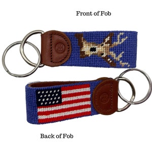 May include: A blue needlepoint keychain with a brown leather backing and silver key rings. The front features a deer design, while the back displays an American flag. The leather backing has an embossed logo.