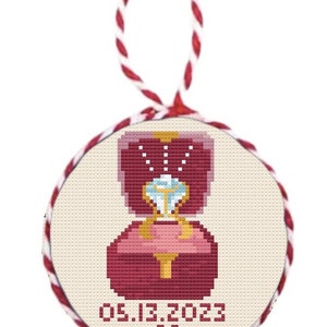 May include: A round, cream-coloured ornament with a red and gold ring box and a diamond ring. The date 05.13.2023 and a red heart are printed below the ring box.