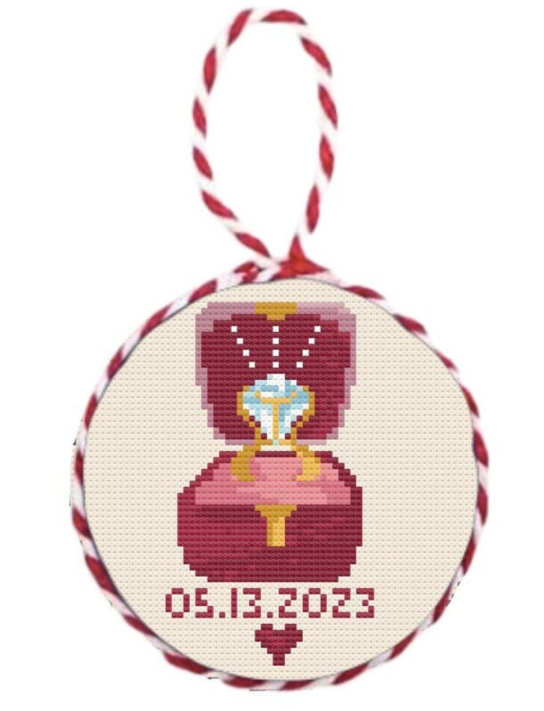 May include: A round, cream-coloured ornament with a red and gold ring box and a diamond ring. The date 05.13.2023 and a red heart are printed below the ring box.