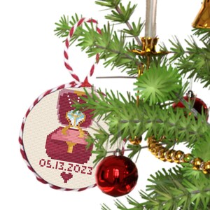 May include: A red and white Christmas bauble with a cross-stitch design of a ring box with a diamond ring inside. The bauble has the date "05.13.2023" on it.