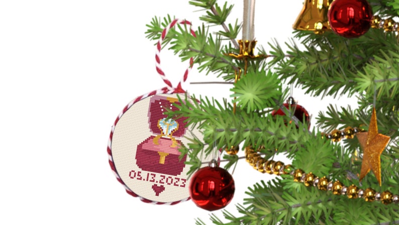 May include: A red and white Christmas bauble with a cross-stitch design of a ring box with a diamond ring inside. The bauble has the date "05.13.2023" on it.