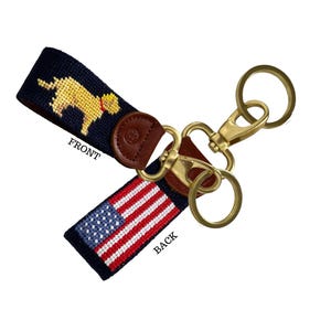 May include: Keychains with a navy blue strap featuring a yellow dog design and the American flag. The keychain has a leather accent and gold-colored metal hardware. The word "FRONT" is visible on the dog side, and "BACK" on the flag side.