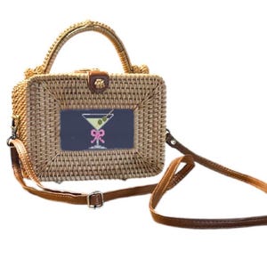 Puede incluir: Bolso de mano de ratán tejido con asa superior y bandolera de cuero marrón desmontable. El bolso rectangular presenta un parche bordado azul marino con un diseño de copa de martini y un lazo rosa. El bolso tiene un cierre de cuero marrón.