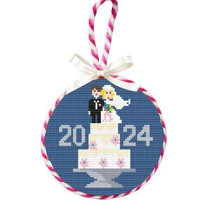 May include: A round, cross-stitched ornament with a blue background and a white and pink braided edge. The ornament features a wedding cake with a bride and groom topper and the year "2024" in white. A white ribbon and pink and white cord are attached.