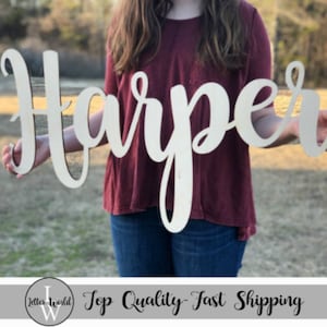May include: A white wooden sign with the name "Harper" written in a cursive font. The sign is being held by a person wearing a maroon shirt and blue jeans.