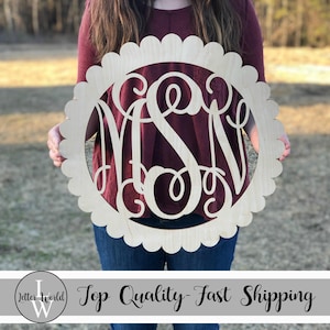 May include: A wooden monogram sign with a scalloped edge, featuring the letters "MSN" in a cursive font. The sign is held by a person wearing a maroon shirt and blue jeans.
