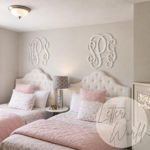 May include: Two white wooden monogram wall art pieces featuring intricate script lettering mounted above two tufted headboards. The beds are dressed in pink quilted coverlets and floral patterned pillows, with a central lamp on a small table.
