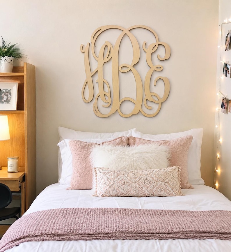 May include: A large, light-colored wooden wall hanging featuring an intricate, interconnected three-letter monogram design with cursive, swirling calligraphy. The piece is mounted centrally on a plain wall above a bed with layered bedding.