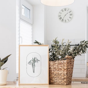 May include: A framed botanical print featuring a line drawing of snowdrop flowers in a minimalist style. The artwork is set against a white background and framed in light wood. A woven basket with greenery and a potted plant are also visible.