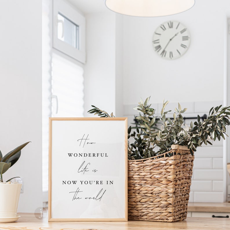 Pode incluir: Uma impress&atilde;o emoldurada com fundo branco e texto preto que diz "How wonderful life is now you're in the world". A impress&atilde;o est&aacute; sobre uma mesa de madeira com uma cesta de vime e uma planta em vaso.
