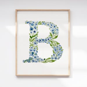 May include: A framed watercolor print featuring the letter "B" composed of blue flowers and green leaves. The artwork is set against a white background and framed in a light wood. The floral design gives a botanical theme.
