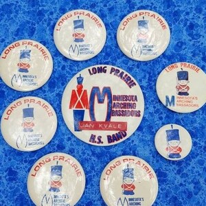 May include: A collection of vintage Long Prairie Minnesota Marching Bassadors band buttons. The buttons are white with red, blue, and black text and graphics. The buttons are arranged on a blue patterned surface.