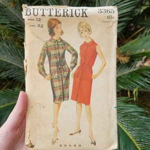 May include: Vintage Butterick sewing pattern envelope featuring illustrations of two dresses. One is a long-sleeved plaid dress with a tie waist, and the other is a sleeveless red dress. The envelope is tan with the text "Butterick" and the number "3365".