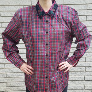 May include: A long-sleeved plaid shirt in red, blue, and green. The shirt has a dark collar with embroidered details and black buttons down the front. The sleeves are cuffed at the wrists.