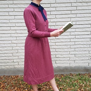 May include: A vintage-style, long-sleeved dress in a deep rose color. The dress features a matching jacket with a navy blue collar and bow. The outfit is paired with a book. The dress is knee-length and has a classic, timeless design.