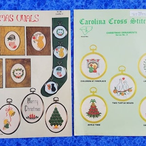 May include: Two vintage Christmas cross-stitch pattern sheets. One sheet features oval designs with cats, a cardinal, and the text "Merry Christmas." The other sheet, from Carolina Cross Stitch, Inc., showcases Christmas ornaments with various designs.