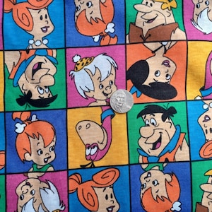May include: A cotton fabric featuring a repeating grid pattern of The Flintstones characters including Fred, Wilma, Pebbles, Bamm-Bamm, and Dino in vibrant colors. A silver-colored coin is placed on the fabric for scale, measuring 0.75 inches in diameter.