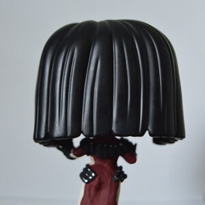 以下が含まれることがあります： 黒と赤のビニール製のフィギュアで、大きな黒い頭と赤と黒の衣装を着ています。フィギュアは木製の表面に立っています。