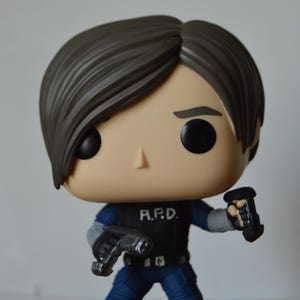 May include: A vinyl collectible figure featuring a character with brown hair, wearing a black vest with the white text R.P.D. printed on the chest, a blue shirt, blue pants, and black boots. The figure holds a black handgun in each hand.