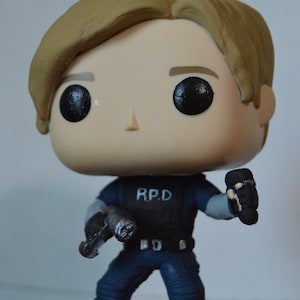 May include: A pop vinyl figure of a man with brown hair wearing a blue uniform with the letters "R.P.D." on the chest. He is holding a gun in one hand and a small object in the other.