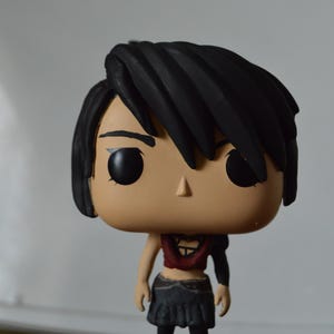Könnte beinhalten: Eine handbemalte, benutzerdefinierte Pop-Vinylfigur einer weiblichen Figur mit schwarzen Haaren, einem roten Oberteil, einem schwarzen Rock und schwarzen Stiefeln. Die Figur steht auf einem durchsichtigen Plastiksockel.