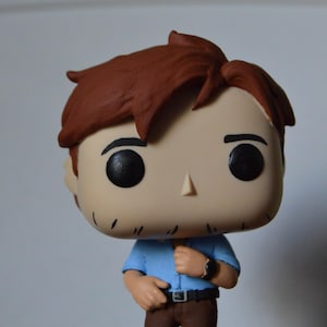 Puede incluir: Una figura de vinilo Funko Pop de un hombre con cabello castaño, que lleva una camisa azul, pantalones marrones y un reloj negro. La figura tiene ojos negros grandes y una nariz pequeña. La figura está de pie.