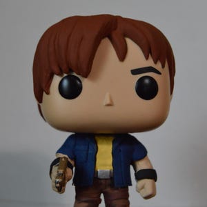 Könnte beinhalten: Eine Vinylfigur eines Charakters mit braunen Haaren, schwarzen Augen und einem gebräunten Teint. Die Figur trägt eine blaue Jacke über einem gelben Hemd, braune Hosen und Stiefel. Sie hält eine goldfarbene Pistole.