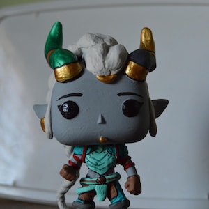 May include: A hand-painted custom pop figure of a grey-skinned character with white hair, green and gold horns, and a teal and brown outfit. The figure is standing on a clear plastic base.
