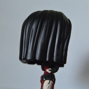 以下が含まれることがあります： 黒と赤でカスタムペイントされた、黒いウィッグをつけたファンコポップフィギュア。フィギュアは木製の表面に立っています。