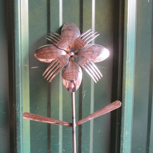 May include: A metal flower sculpture made from spoons and forks. The flower is silver and has six petals. The stem is made from a metal rod and has two leaves.
