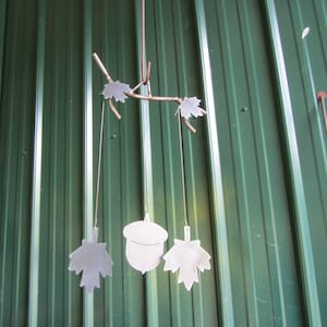 May include: A metal wind chime with a branch design and hanging leaves and an acorn. The chime is hanging from a green metal wall.